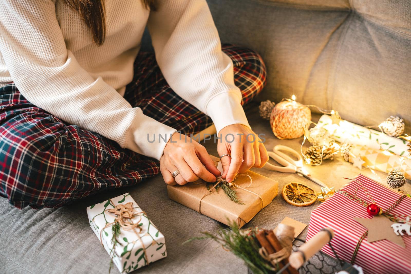 Woman s hands wrapping Christmas gift boxes, close up. Cropped female sit and preparing natural eco presents on couch with decor elements and items. Merry Christmas or New year DIY packing Concept.