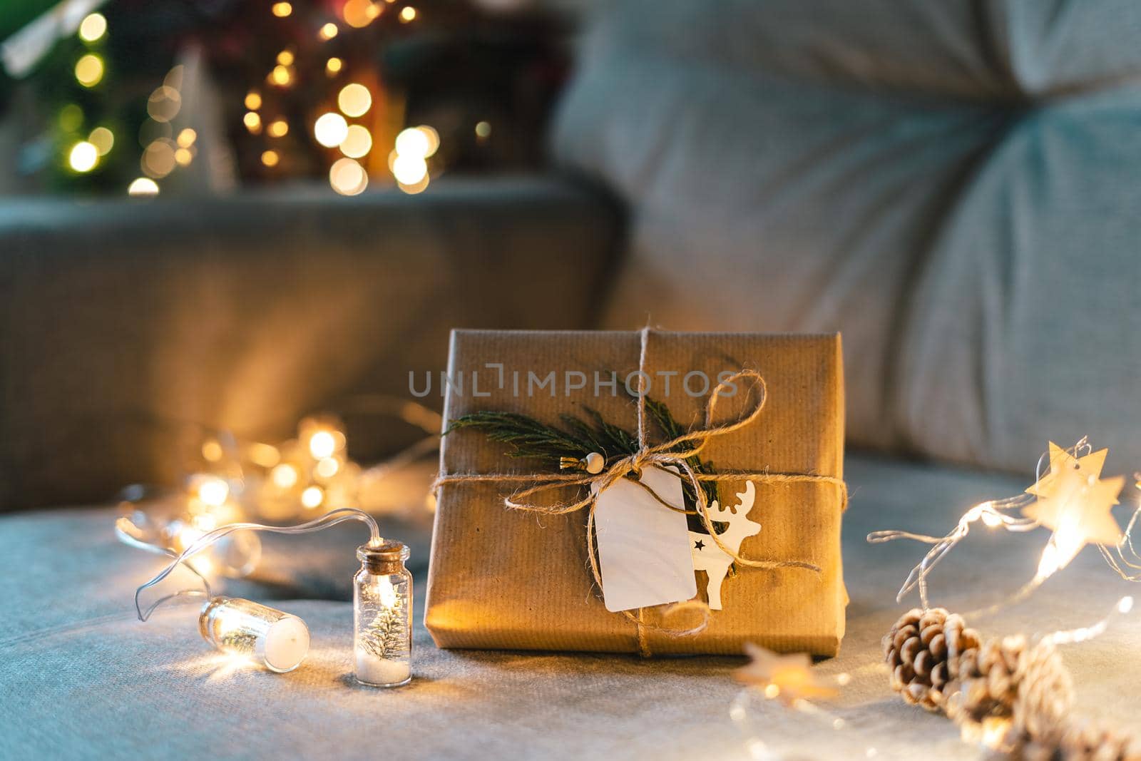 Christmas natural eco gift boxes with empty label tag. Glowing light bokeh, coniferous branch. Winter holidays mock up on gray couch background at cozy home. Zero waste, plastic free. Indoors plants
