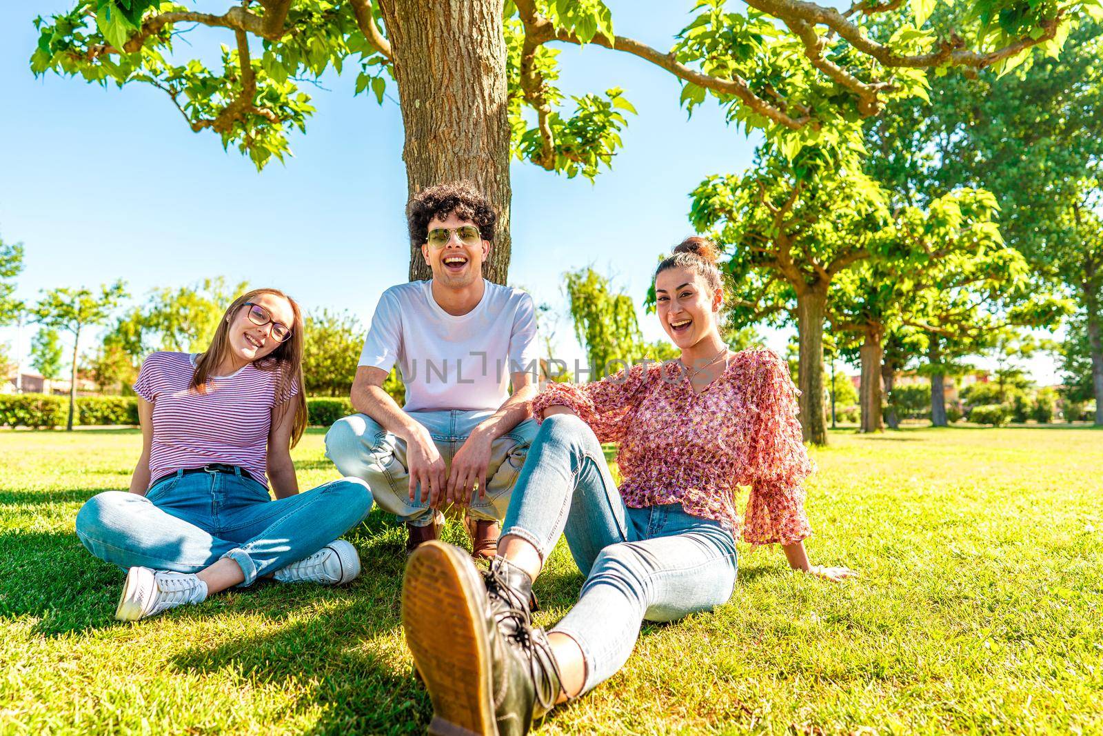 Three young diverse friends resting on green park field smiling looking at camera for a portrait. Gen z students spending time in nature to breaking city life routine. Friendship makes no difference by robbyfontanesi