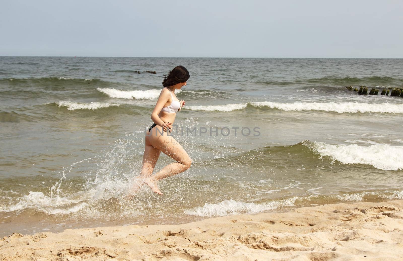 young beautiful brunette girl in a black swimsuit running along the sand of the sea shore on sunny summer day