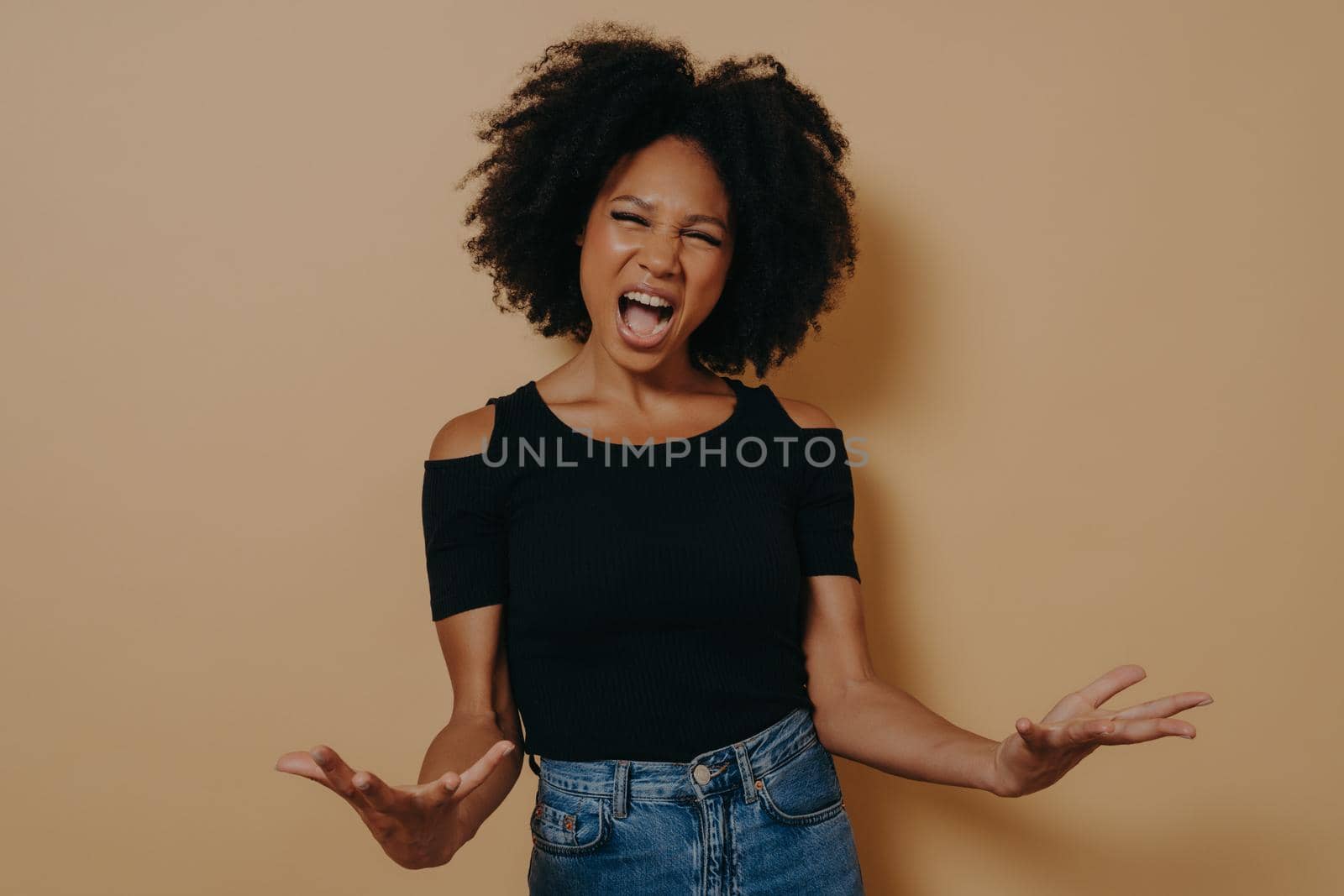 African woman in black t-shirt posing screaming, making palms open, feeling mad, with mouth open wide shouting aloud looking at camera, isolated on beige studio background. Negative emotions concept
