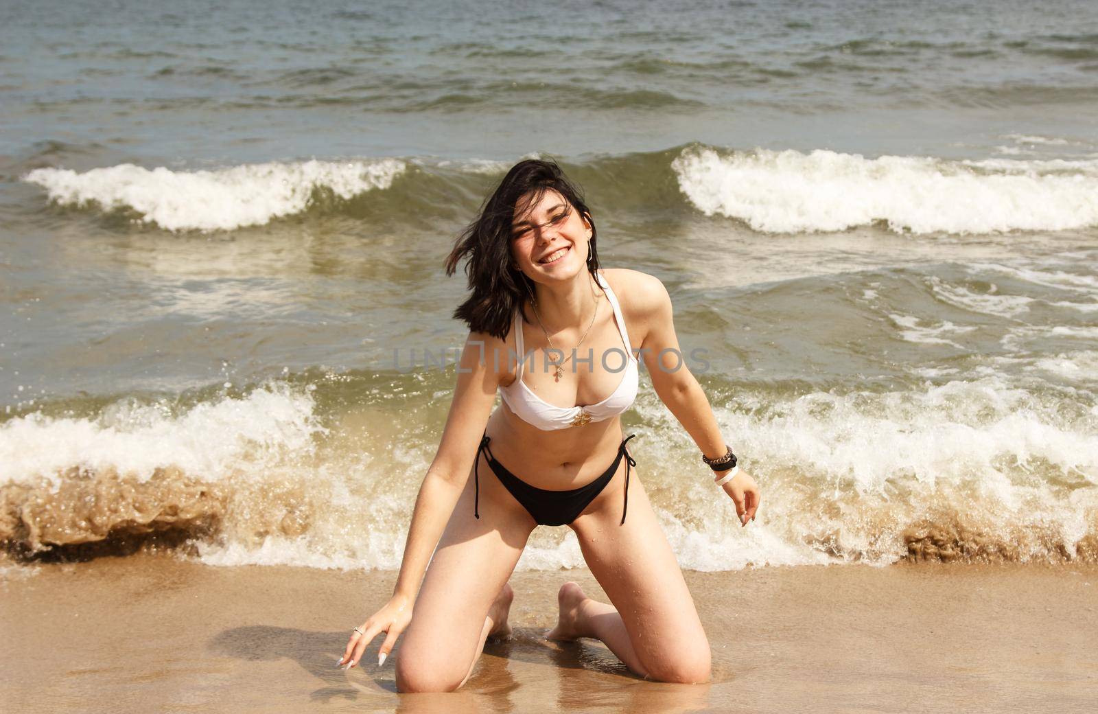 young brunette woman in bikini posing kneeling on the sand by the sea on sunny summer day