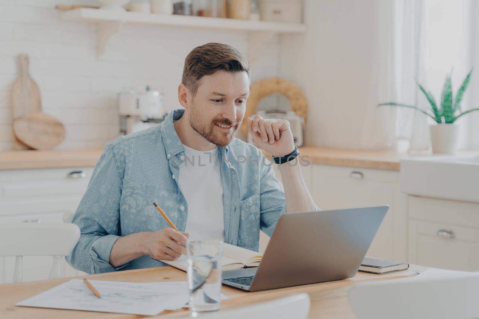 Smiling male employee using laptop at home sitting at kitchen table, looking at screen and making video call or watching webinar, writing notes in notebook. Distance studying and freelance concept