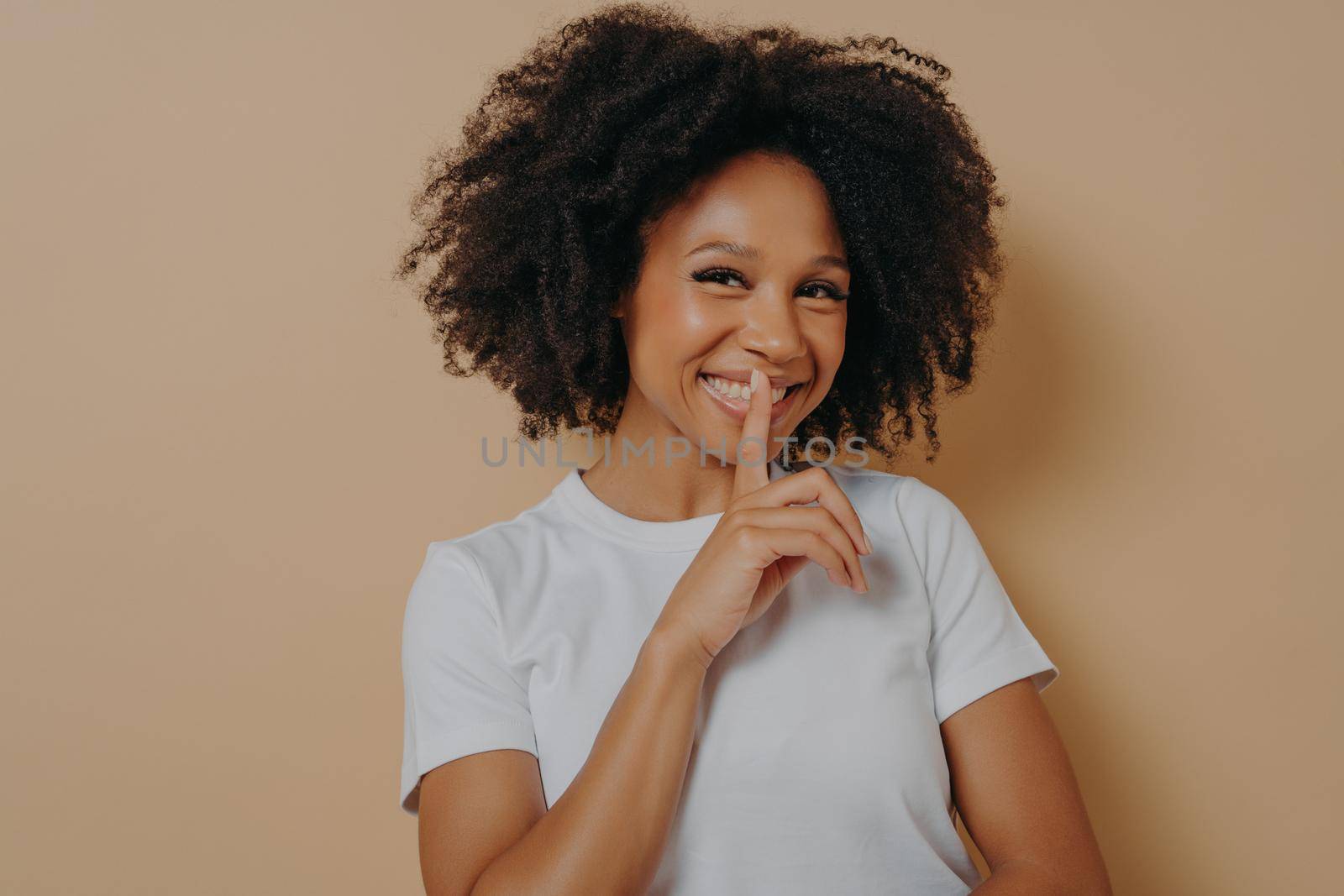 Cheerful smiling afro girl showing shhh sign, keep silence gesture, with index finger near lips, standing over pastel beige background and looking at camera. Positive emotions and body language