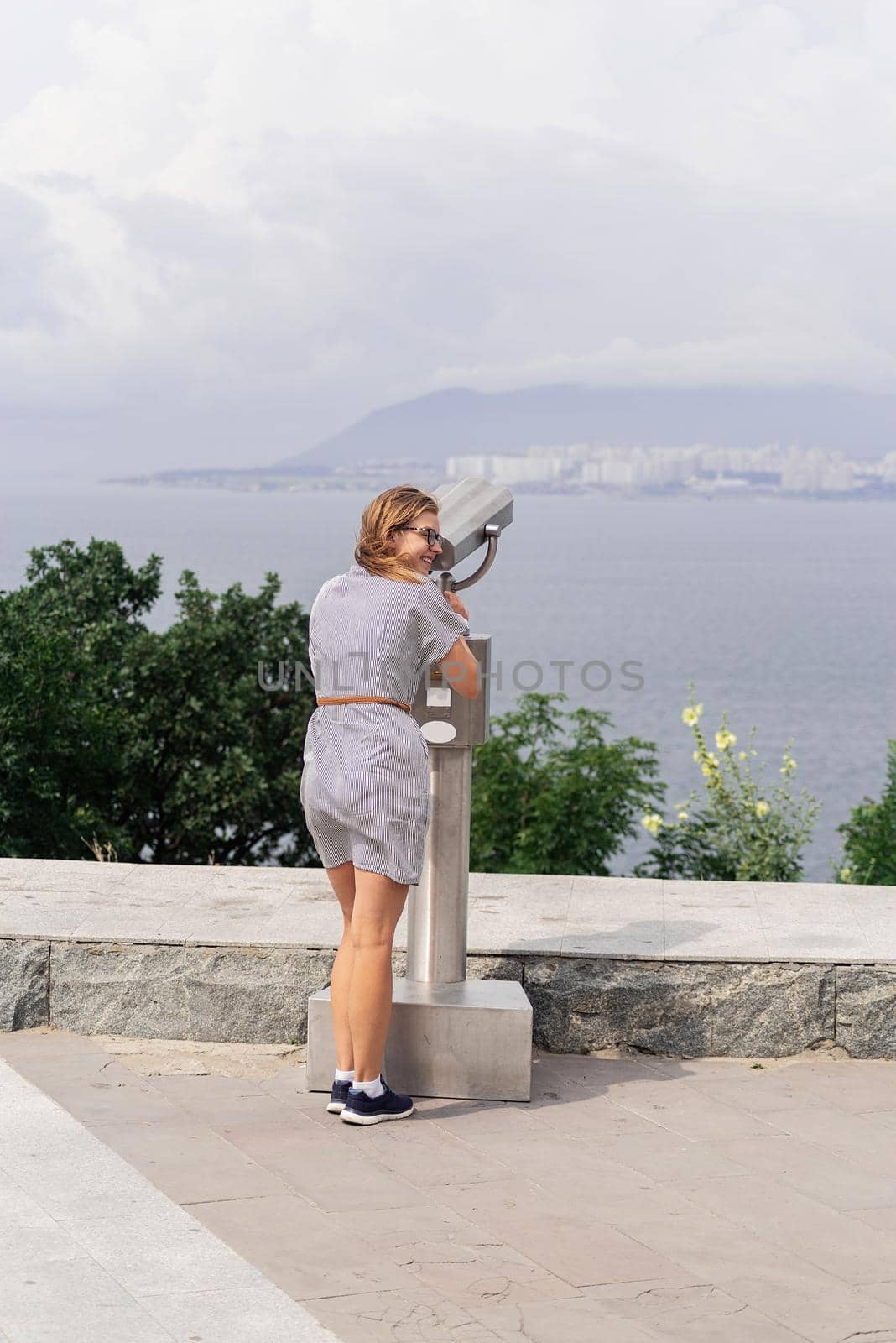 young woman in summer dress watching what is happening with stationary binoculars on a warm summer day on a hill against the background of sea and city