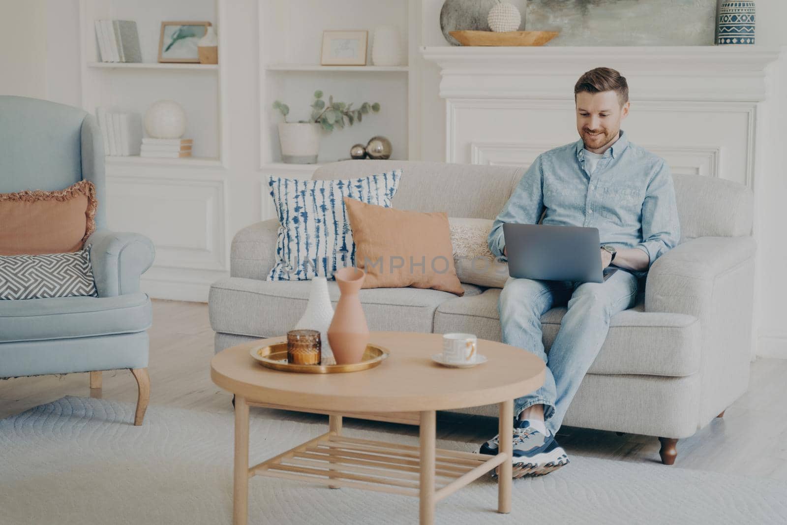 Young man with stubble working as freelancer, using notebook for work, trying to finish commission in time while sitting on comfy small beige couch in modern themed living room