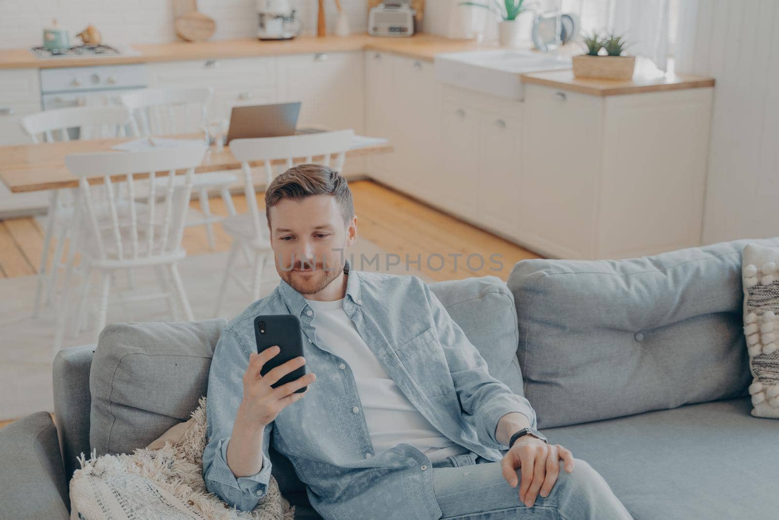 Caucasian young man in domestic casual clothes using smartphone while resting on cozy couch at home, handsome guy sitting on sofa in living room and browsing internet or messaging with friends