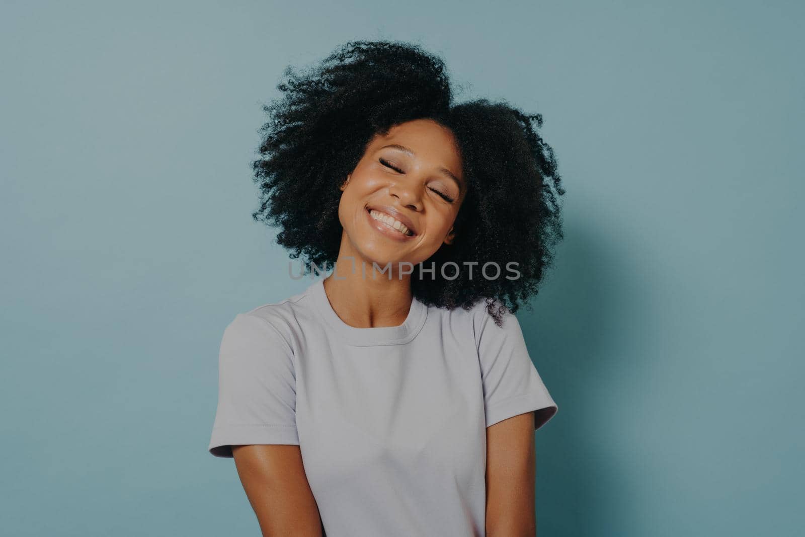 Beautiful overjoyed mixed race woman smiling and looking at camera. Portrait of cheerful young woman with afro hairstyle isolated over pastel blue studio background with copy space