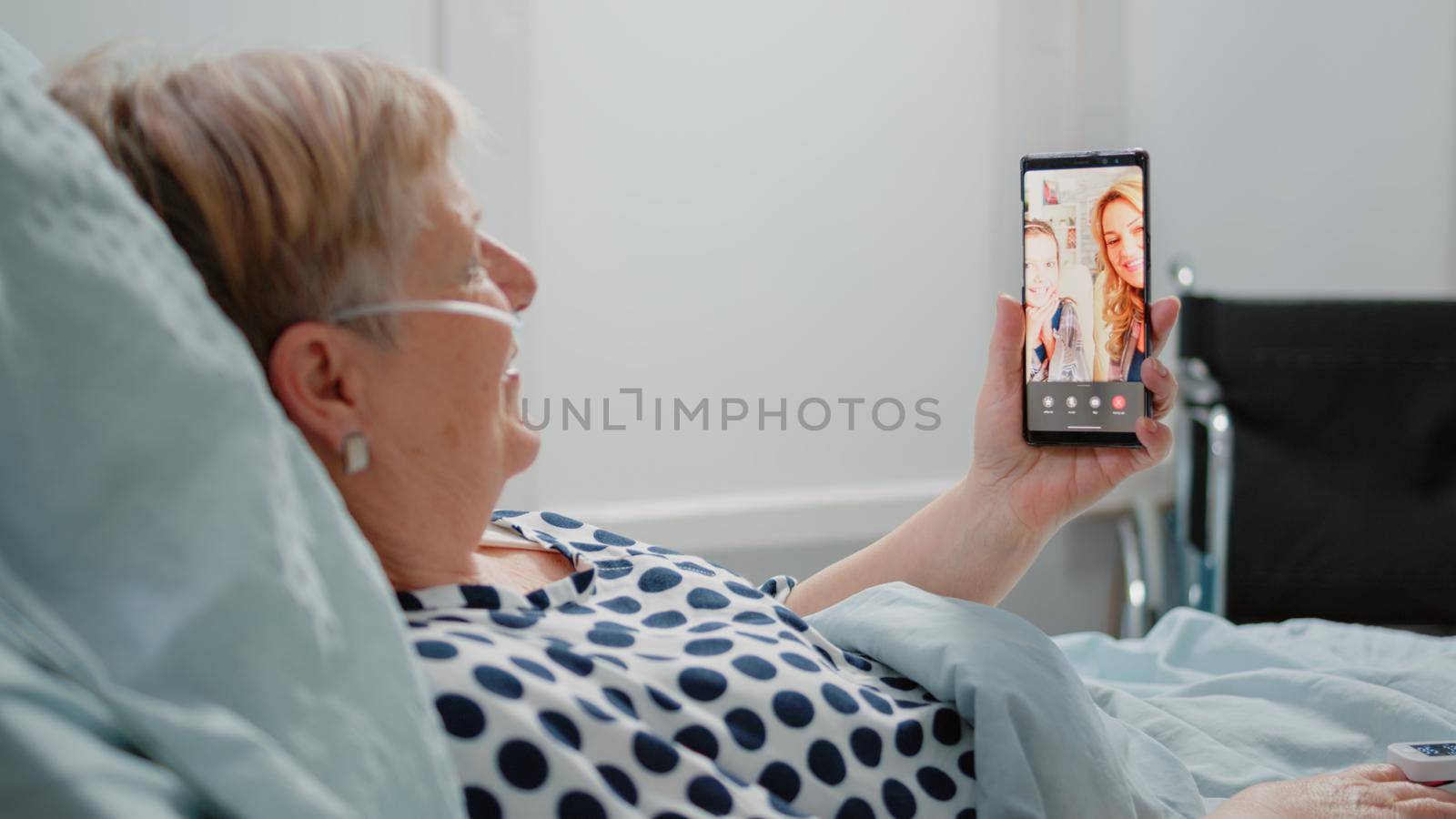 Aged woman holding smartphone with video call for remote communication with niece and daughter. Sick patient using online conference to talk to relatives while laying in hospital ward bed.