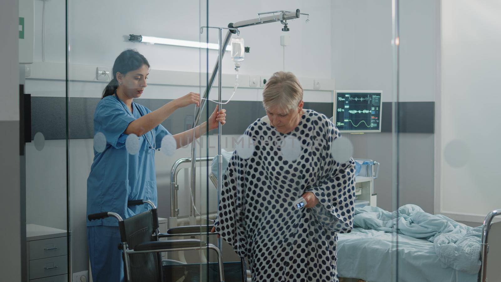 Medical assistant helping sick patient to sit in wheelchair at facility. Nurse giving assistance and support to senior woman with IV drip bag and oximeter for transportation and mobility