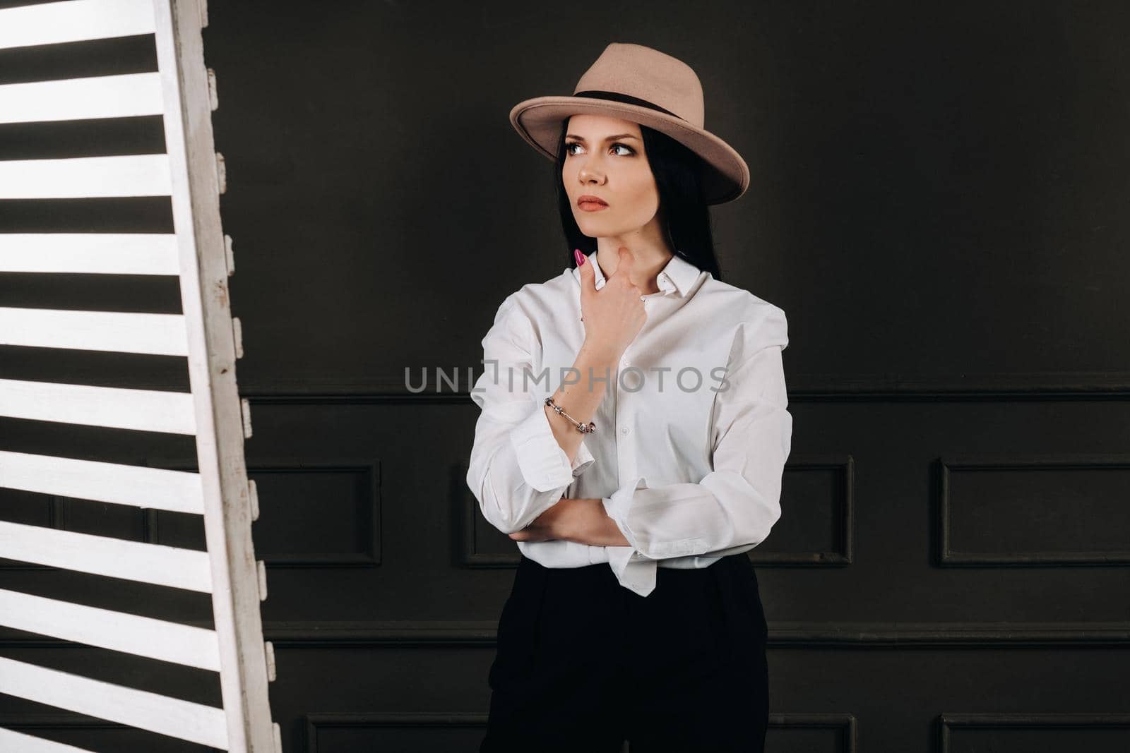 Elegant brunette girl in a white shirt and beige hat on a dark background.