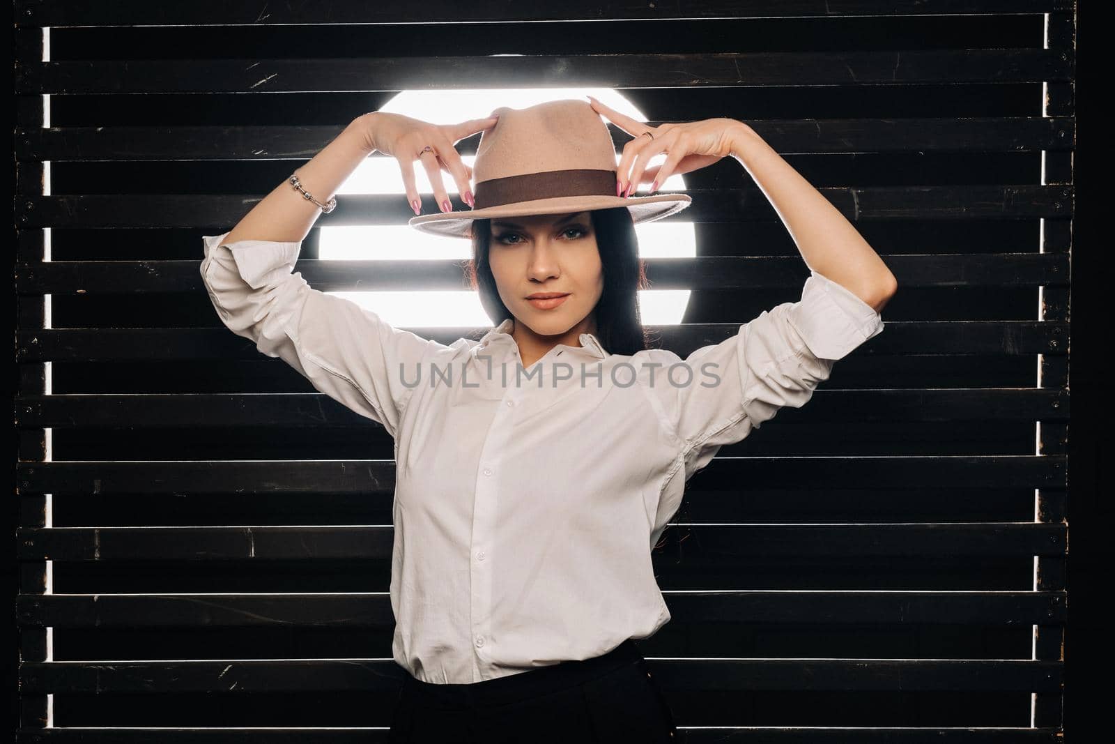 Elegant brunette girl in a white shirt and beige hat on a dark background.