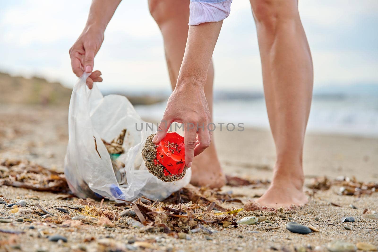 Close-up of a woman's hands picking up plastic trash into a bag on the beach by VH-studio