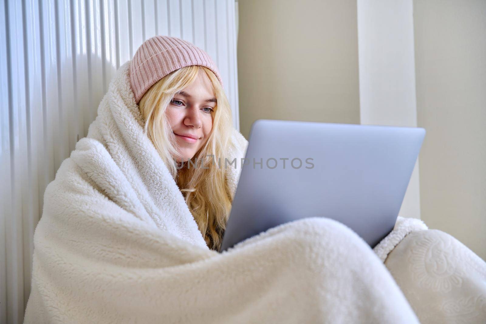 A young female teenager frozen in house in winter cold season, warming up with a warm blanket, hat, central heating radiator, looking at laptop screen, student, having rest