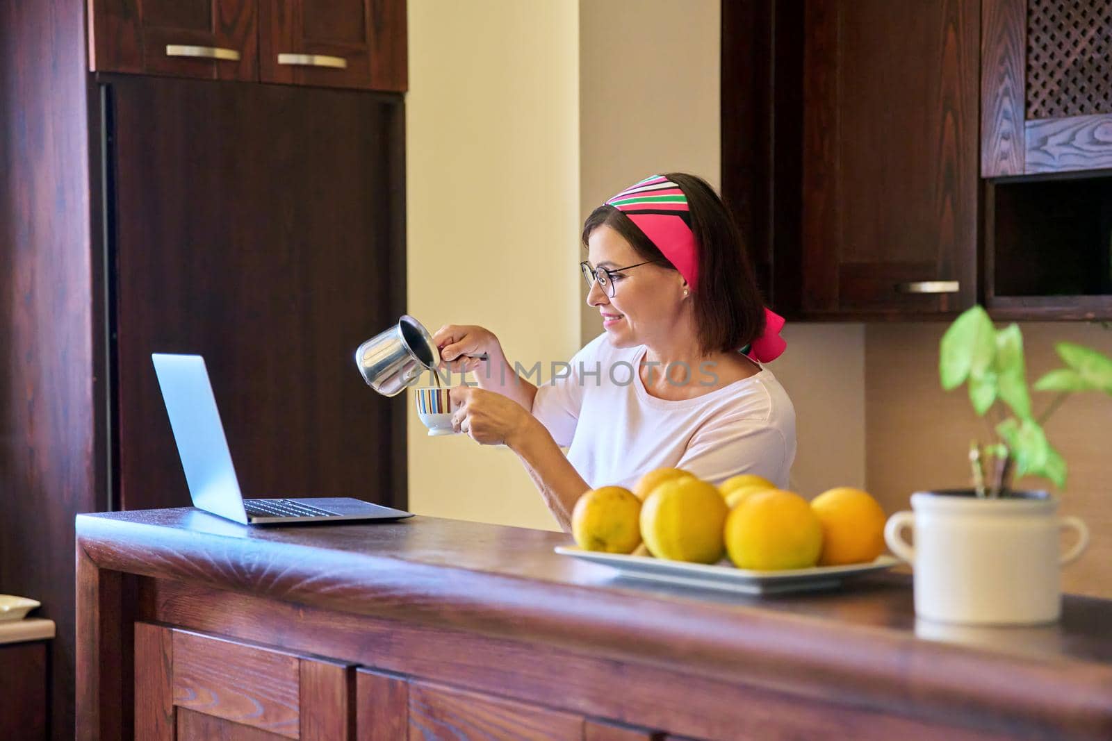 Woman in the kitchen with a cup of coffee looking into the laptop screen. Middle aged woman using video call for meeting and morning coffee. Technology, lifestyle, home, mature people concept