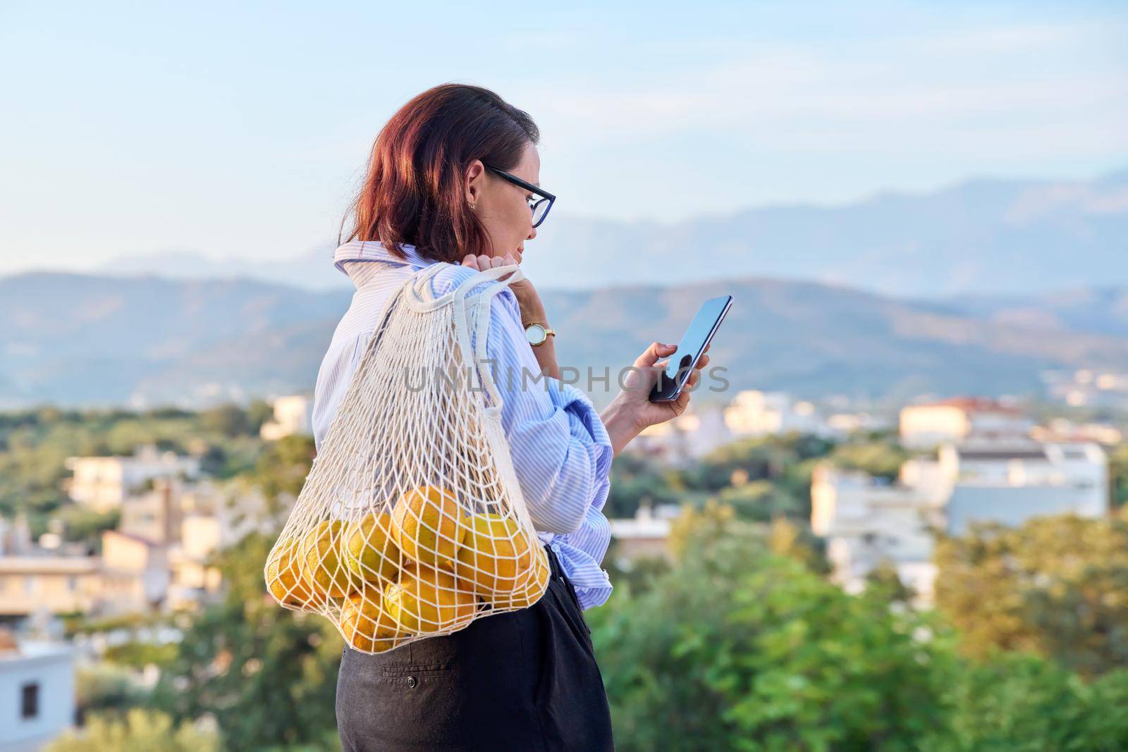 Portrait of beautiful middle-aged woman with smartphone trending grid of oranges outdoors. Female with phone and eco mesh shopping bag with organic oranges, mountains village sunset background, trends