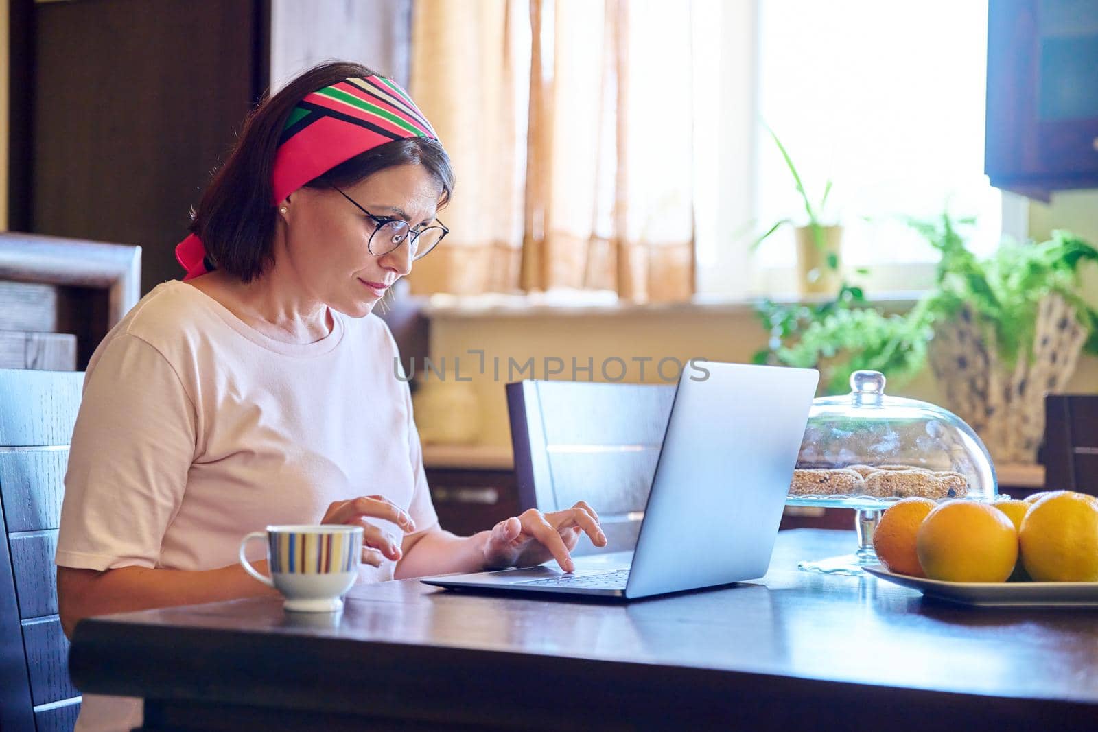 Middle-aged woman sitting at home in kitchen with laptop and cup of coffee. Lifestyle, leisure, technology, work, relaxation, mature people concept