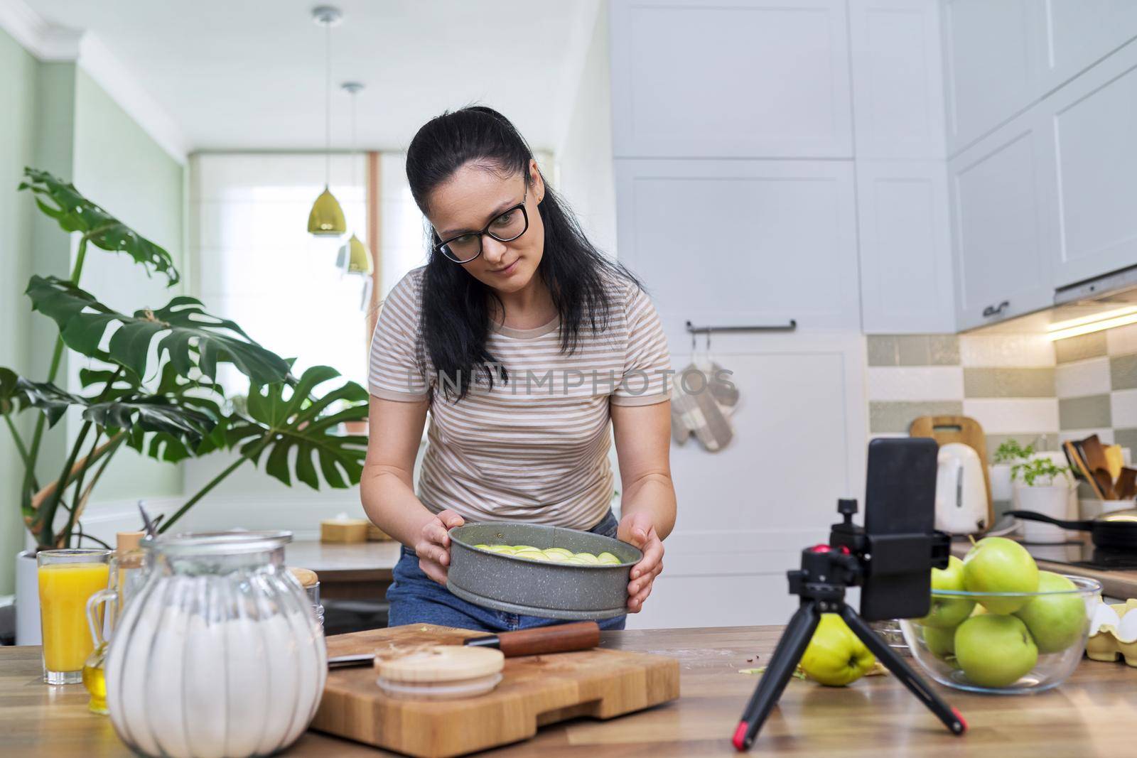 Woman preparing apple pie at home in the kitchen, with a smartphone using a video call by VH-studio