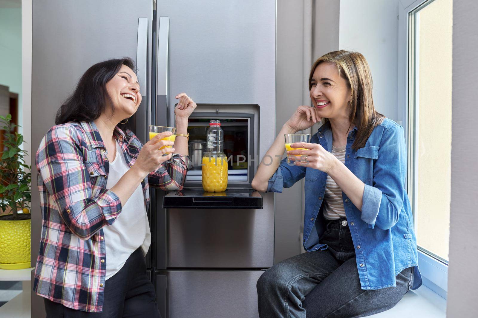Two middle-aged women drinking orange juice. Smiling female friends talking in the kitchen near the refrigerator. People, food, drinks, diet, rest, communication, lifestyle concept