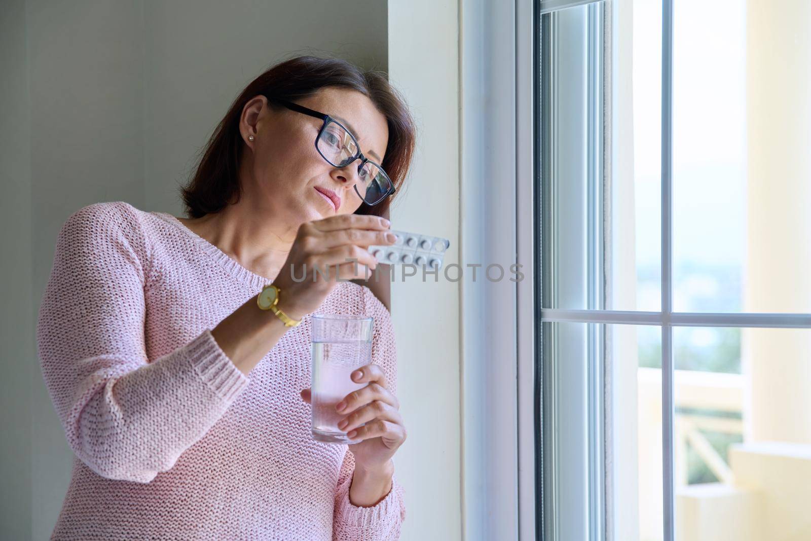 Middle-aged woman with pills and glass of water in her hands, at home by VH-studio