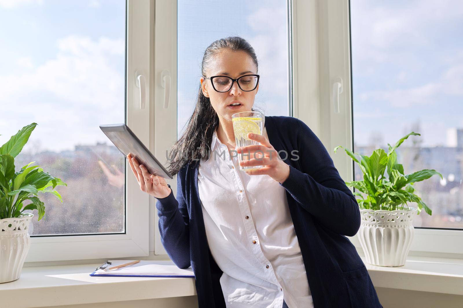 Adult woman drinks water with lemon, female in glasses with digital tablet, at home near window