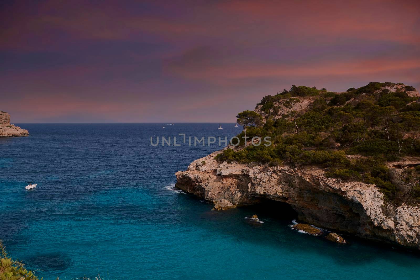 Cliff on turquoise beach with orange sunrise or sunset sky. Balearic Islands, Spain, Mediterranean Sea, unmanned, Sailboats