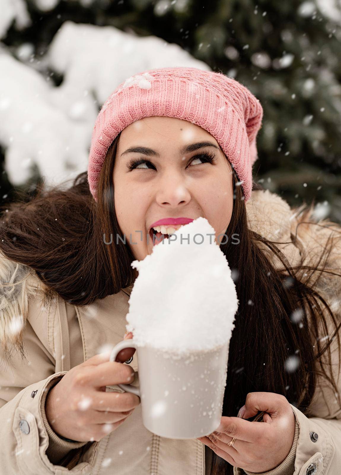 woman in warm winter clothes standing by the big christmas tree outdoors and licking snow from the cup by Desperada