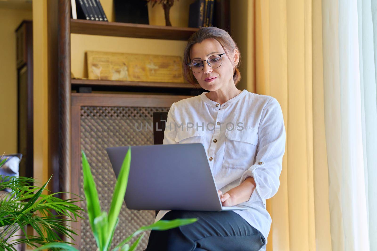 Middle-aged woman psychologist in an office with laptop. Portrait of professional female counselor, psychiatrist, therapist, teacher sitting on chair near window. Technology, medicine, people