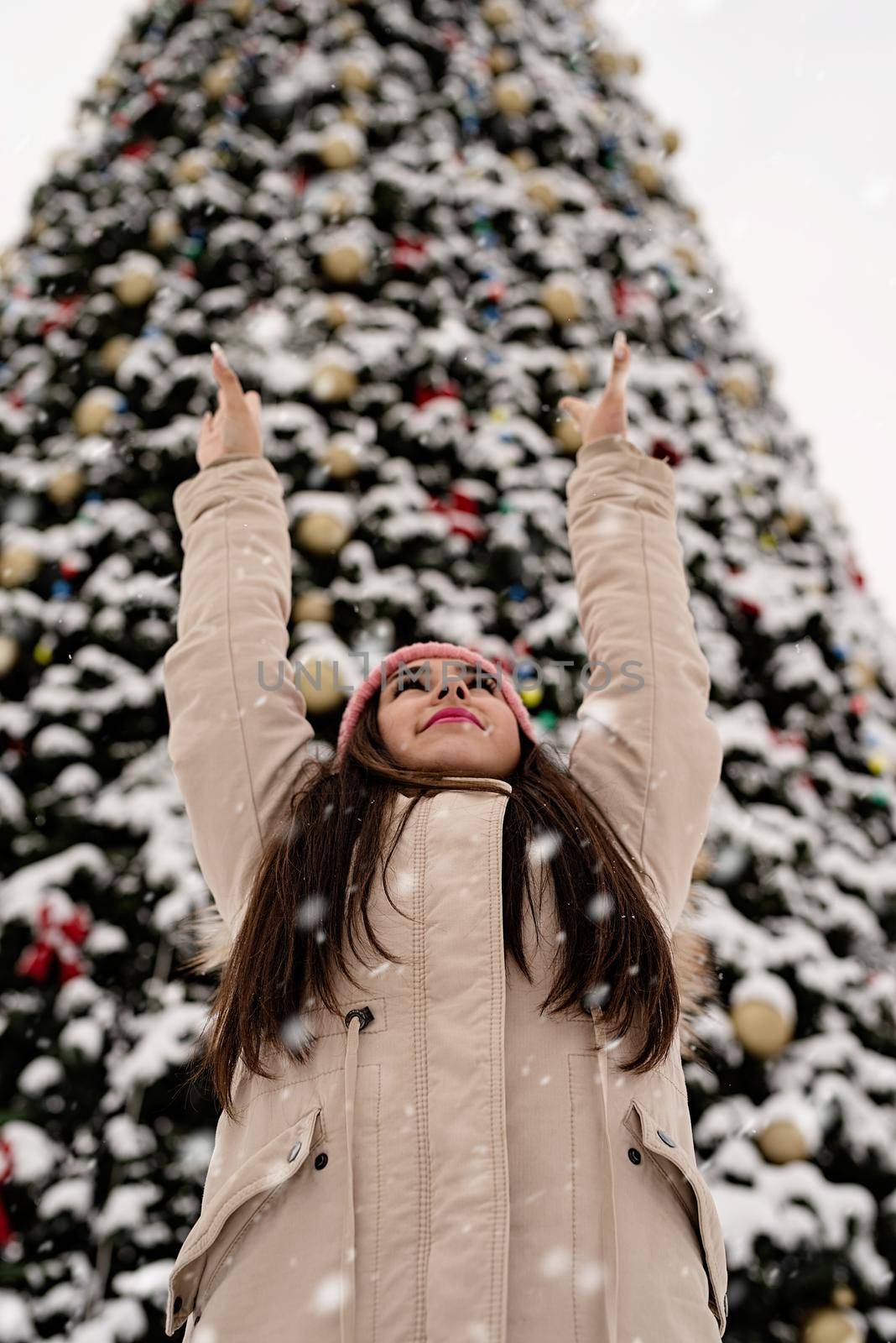 Woman in warm winter clothes standing by the big christmas tree outdoors, arms up, snow falling by Desperada