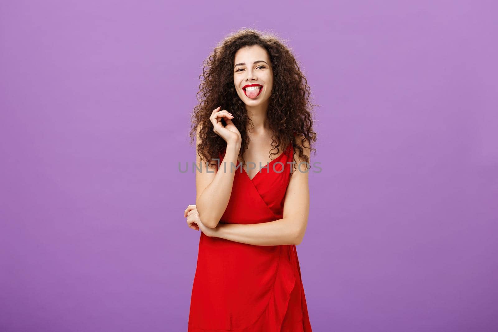 Studio shot of playful and joyful artistic caucasian female student in red evening dress sticking out tongue happily having fun playing with curl standing over purple background careless and carefree.