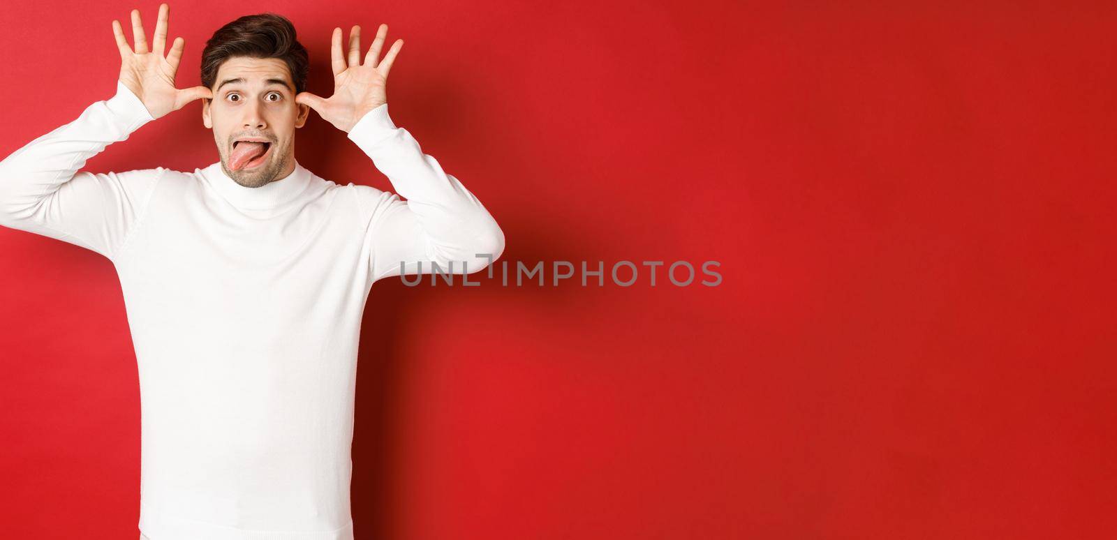 Portrait of funny caucasian guy, showing tongue and making faces, wearing white sweater, standing against red background by Benzoix