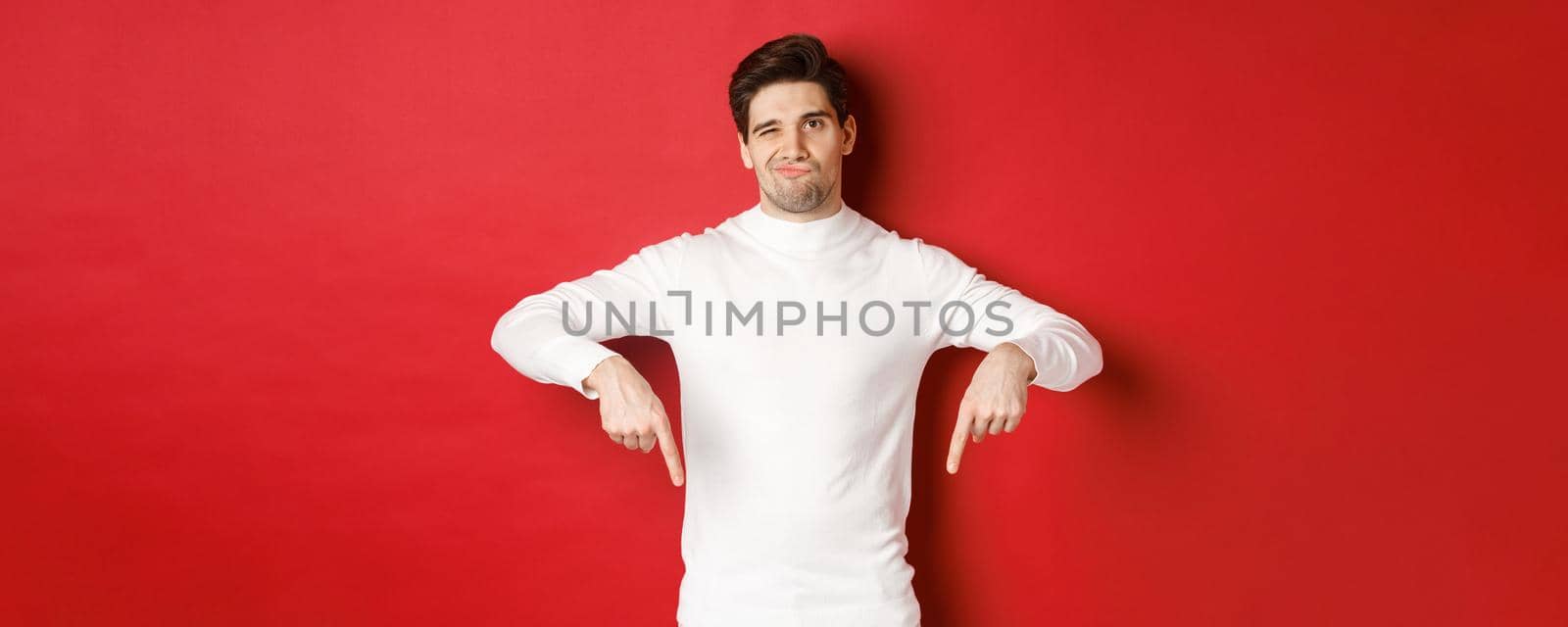 Portrait of attractive, skeptical young man in white sweater, grimacing and pointing fingers down, disagree and dislike something, standing over red background by Benzoix