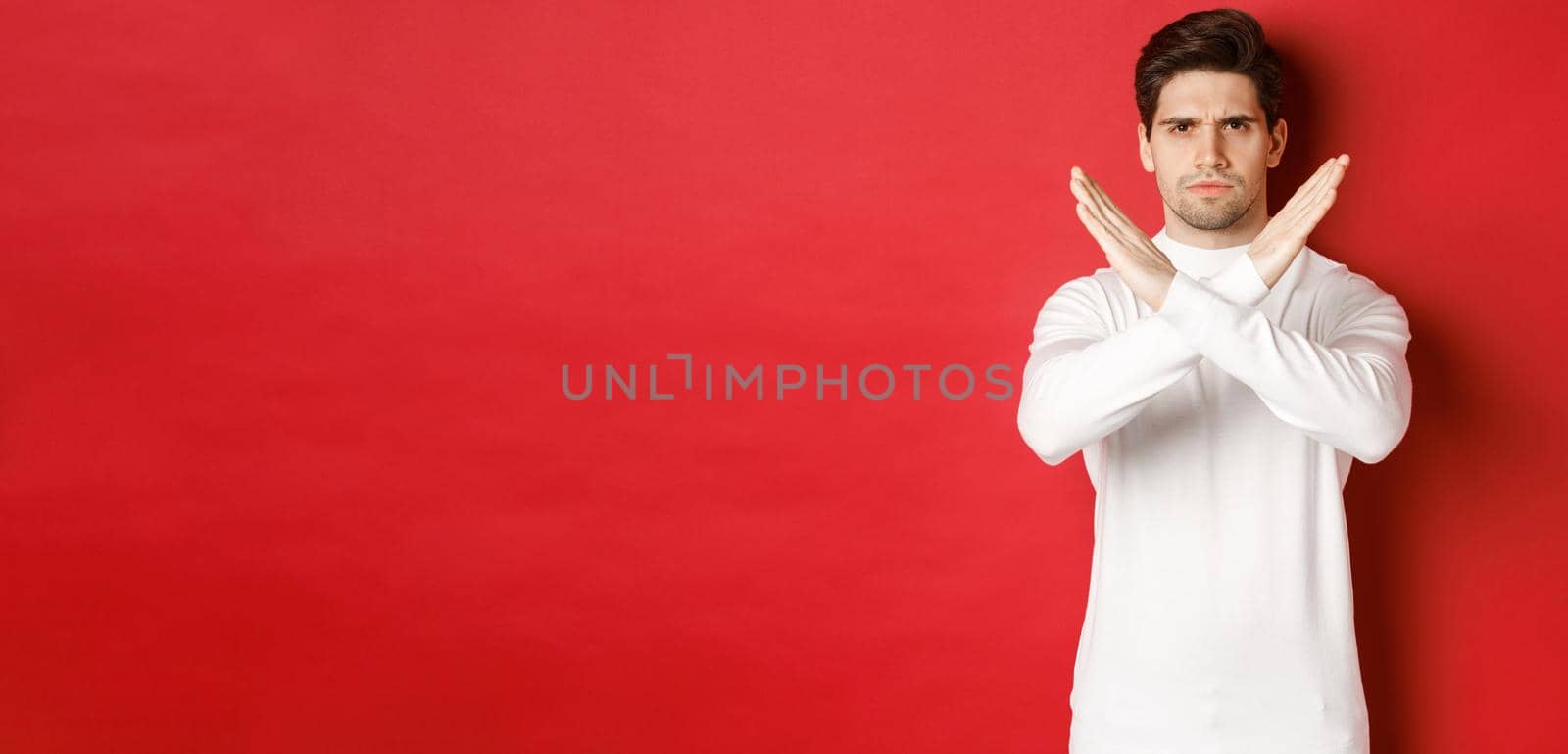 Image of serious and angry man in white sweater, express storng disapproval, showing cross sign to stop something bad, forbid action, standing over red background.