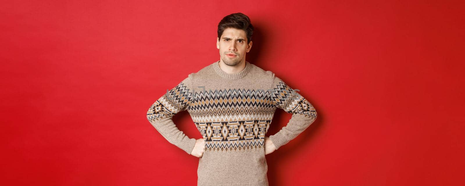 Portrait of doubtful and disappointed man, wearing christmas sweater, frowning and looking displeased at you, standing against red background.