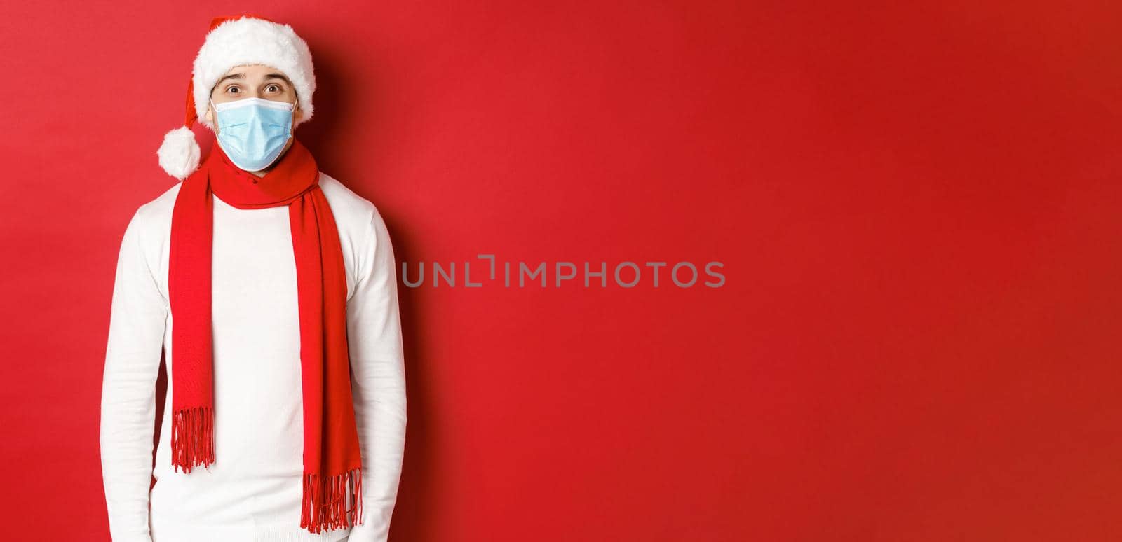 Concept of new year, coronavirus and holidays. Cheerful man celebrating christmas and social distancing, wearing medical mask, santa hat and scarf, standing over red background by Benzoix