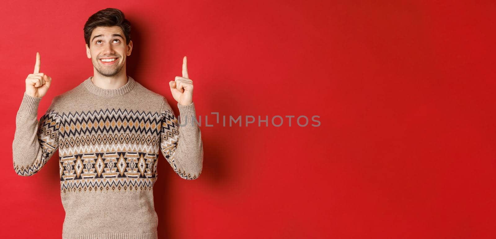 Portrait of dreamy handsome guy thinking about christmas holidays, wearing winter sweater, pointing and looking up at new year promo offer, standing over red background by Benzoix