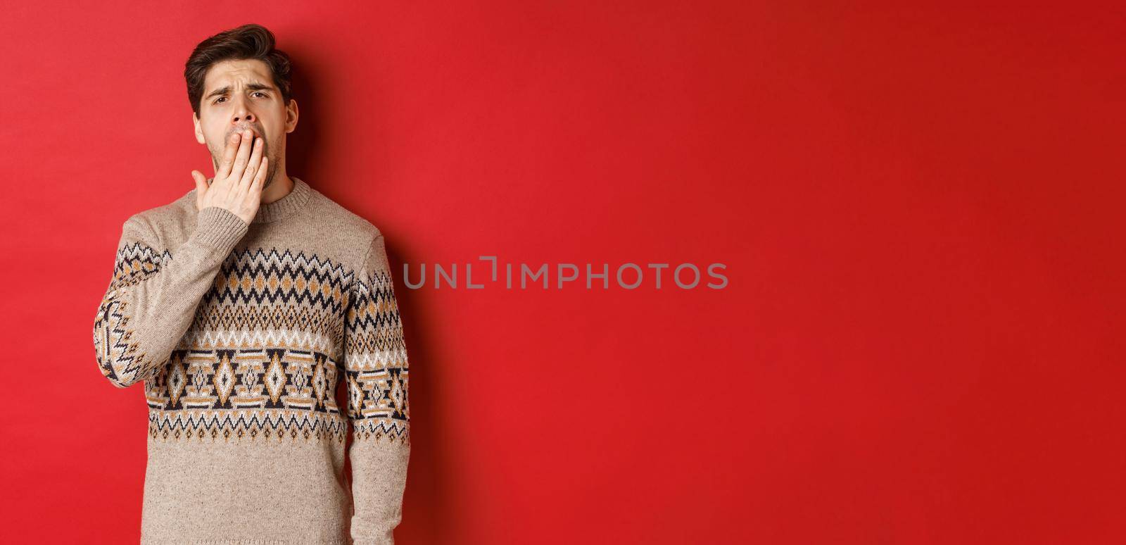 Image of tired or bored handsome man in winter sweater, yawning and covering mouth with hand, standing exhausted against red background.