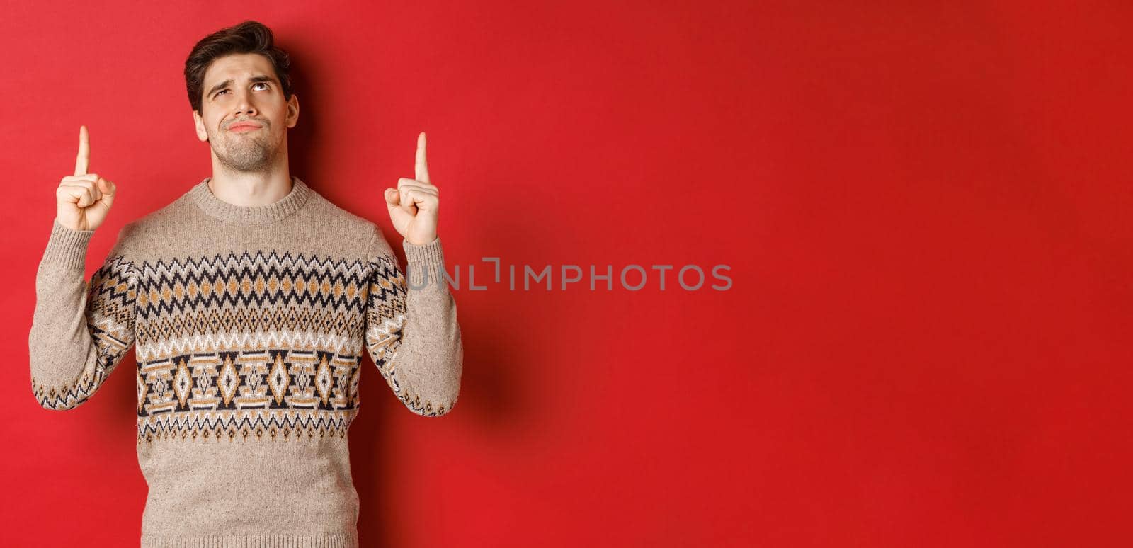 Image of disappointed and skeptical handsome man, wearing christmas sweater, smirk and frowning as looking at something bad, pointing fingers up, standing over red background.