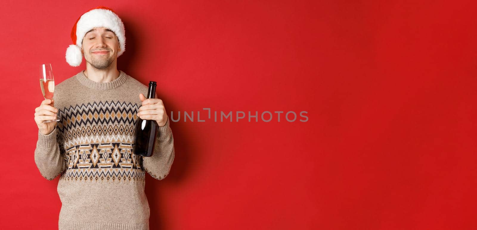 Concept of winter holidays, christmas and celebration. Image of pleased smiling man in santa hat and sweater, drinking on new year, holding bottle of champagne and filled glass, red background.