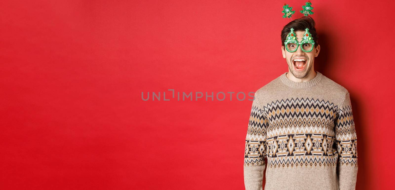Portrait of surprised and excited handsome guy in party glasses and winter sweater, celebrating christmas and having fun, standing against red background.