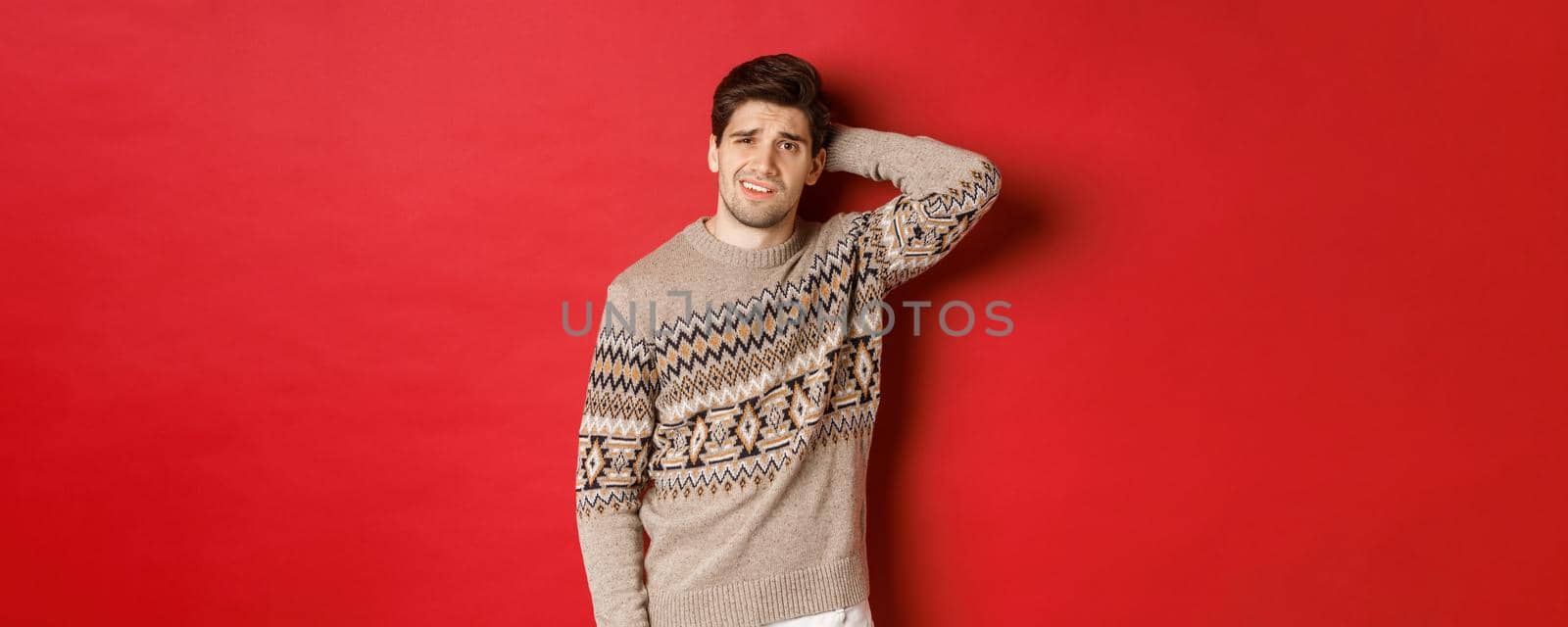 Portrait of confused man in christmas sweater, scratching back of head and looking clueless about new year presents, standing over red background.