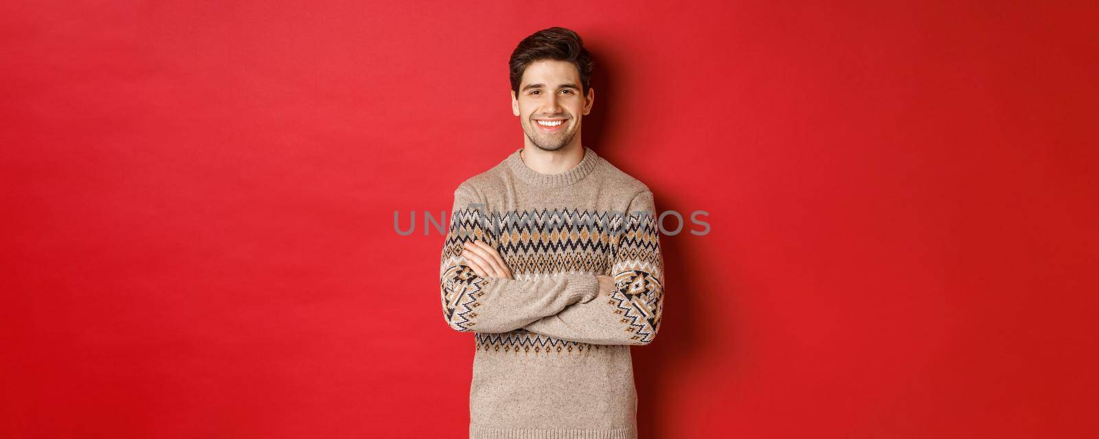 Image of handsome happy guy in christmas sweater, smiling and looking at camera, celebrating xmas holidays, standing over red background.