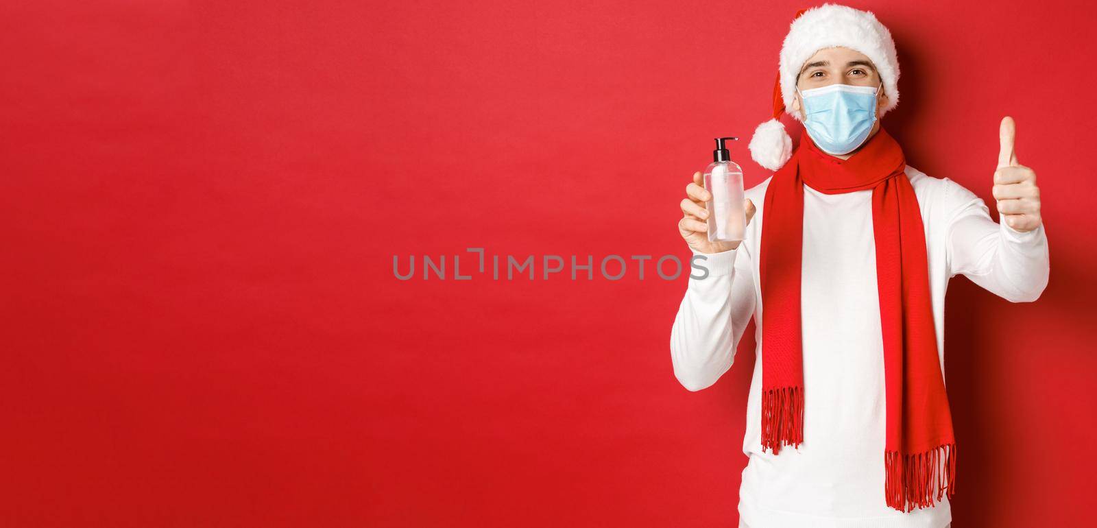 Concept of covid-19, christmas and holidays during pandemic. Happy and satisfied man in santa hat and medical mask, showing thumb-up in approval and recommending hand sanitizer by Benzoix