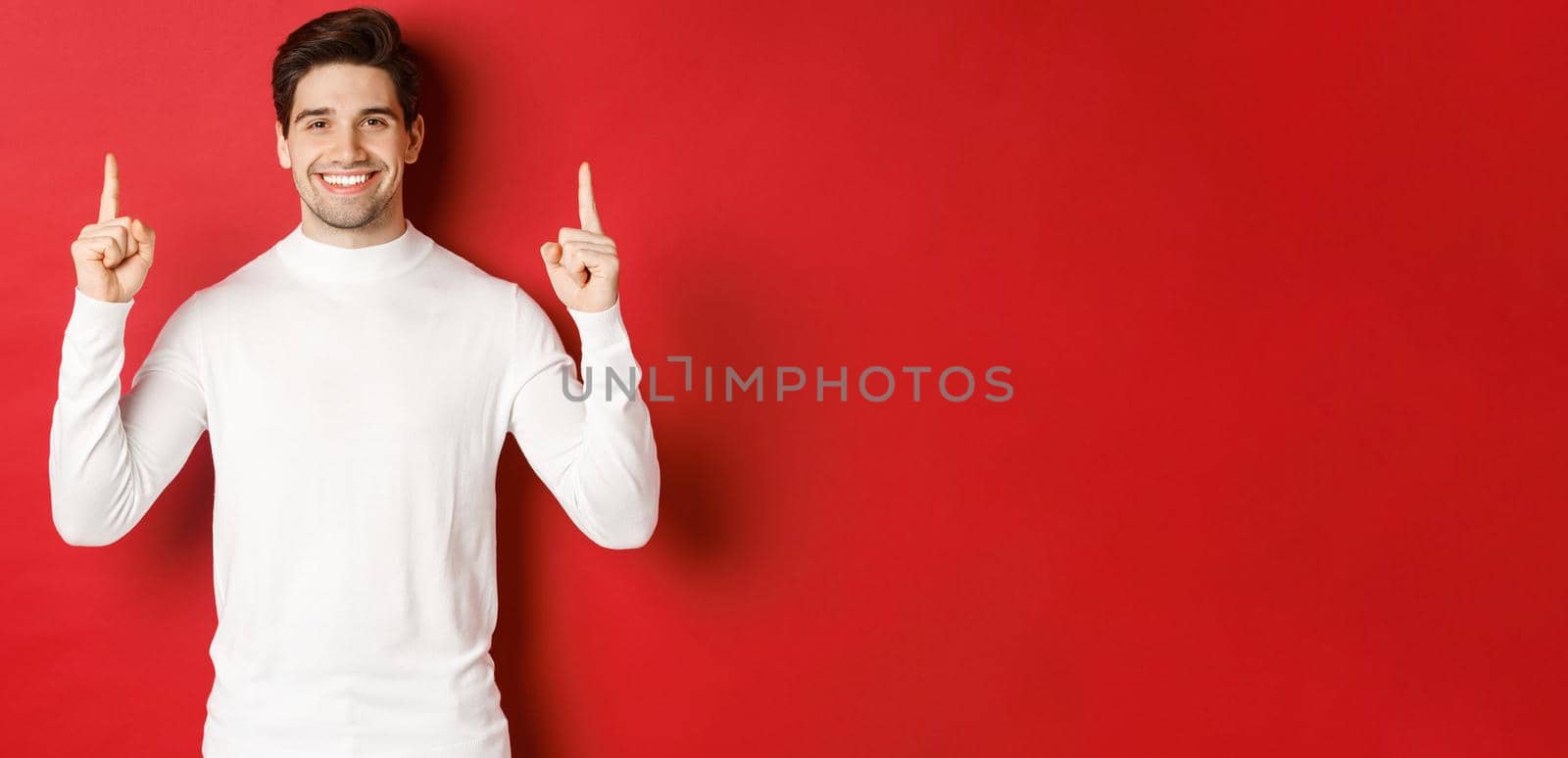 Concept of winter holidays. Good-looking young man with bristle, wearing white sweater, showing christmas advertisement on copy space, pointing fingers up and smiling, red background by Benzoix