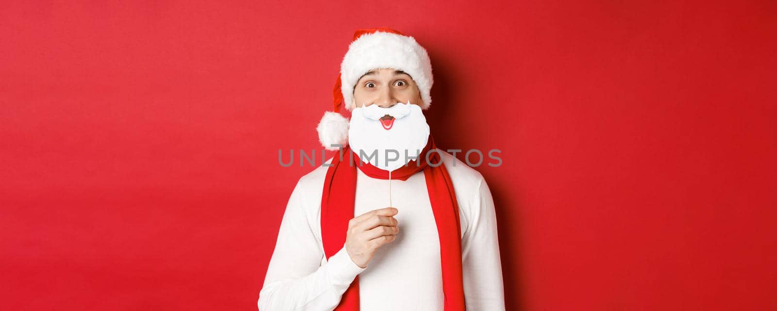 Concept of christmas, winter holidays and celebration. Portrait of funny man in santa hat, holding beard mask, enjoying new year party, standing over red background.