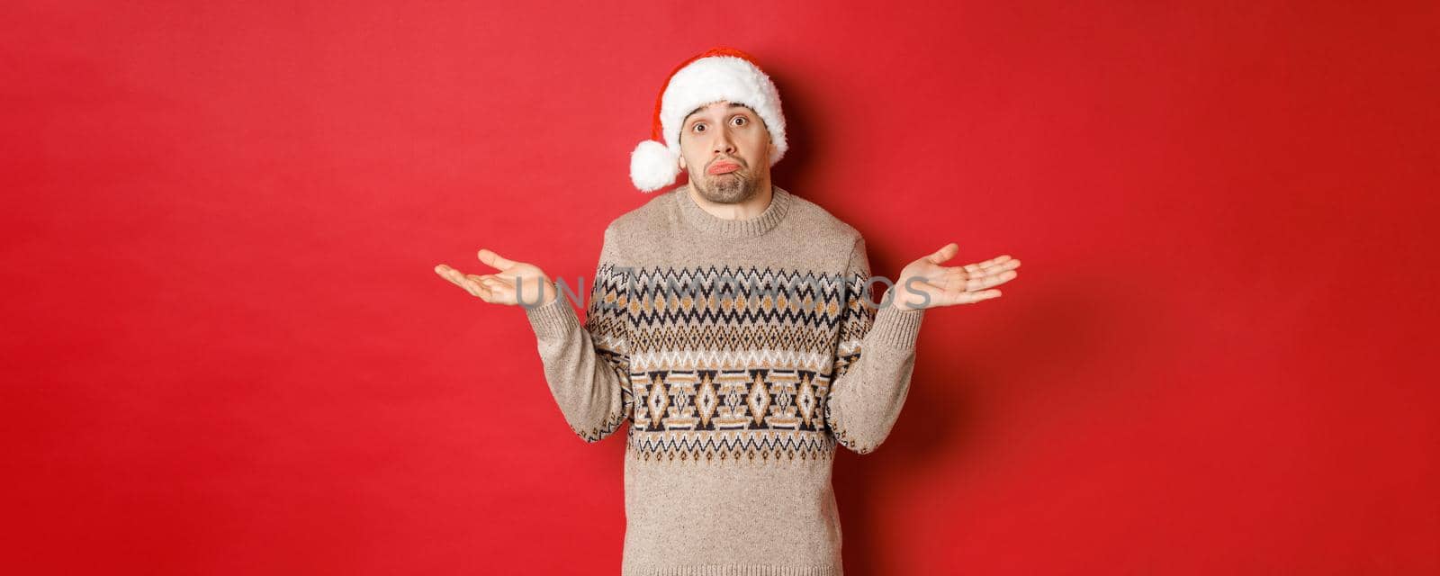 Image of clueless young man in sweater and santa hat, shrugging and looking unaware, dont know what to buy for new year holidays, standing over red background by Benzoix