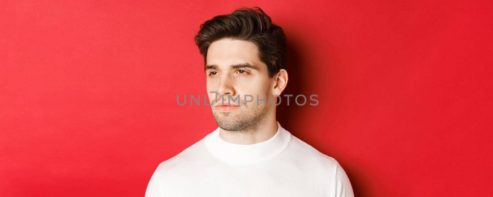 Close-up of serious and thoughtful man in white sweater, looking left, standing against red background.