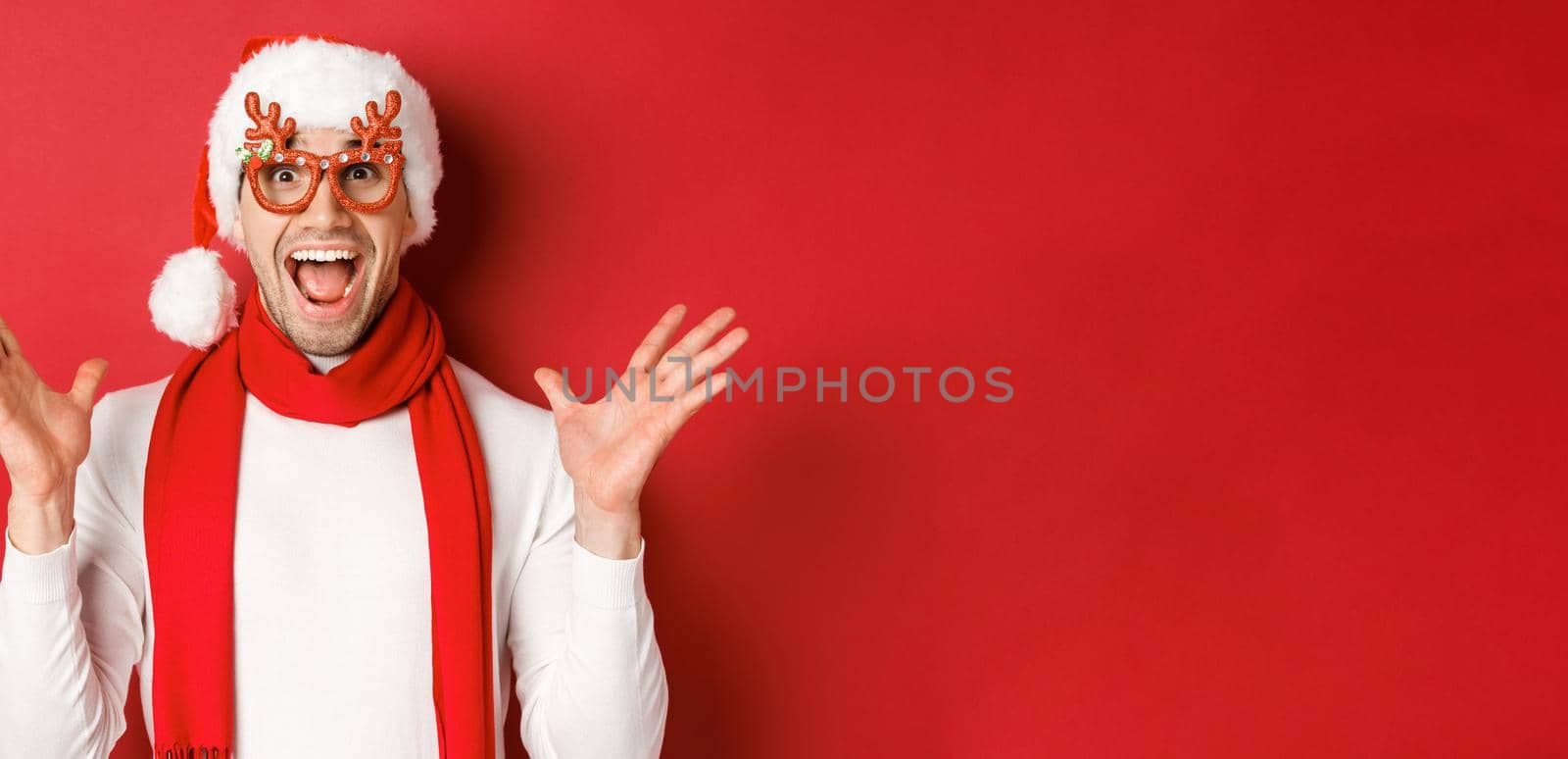 Concept of christmas, winter holidays and celebration. Image of surprised and happy man looking amazed, wearing party glasses and enjoying new year, standing over red background by Benzoix