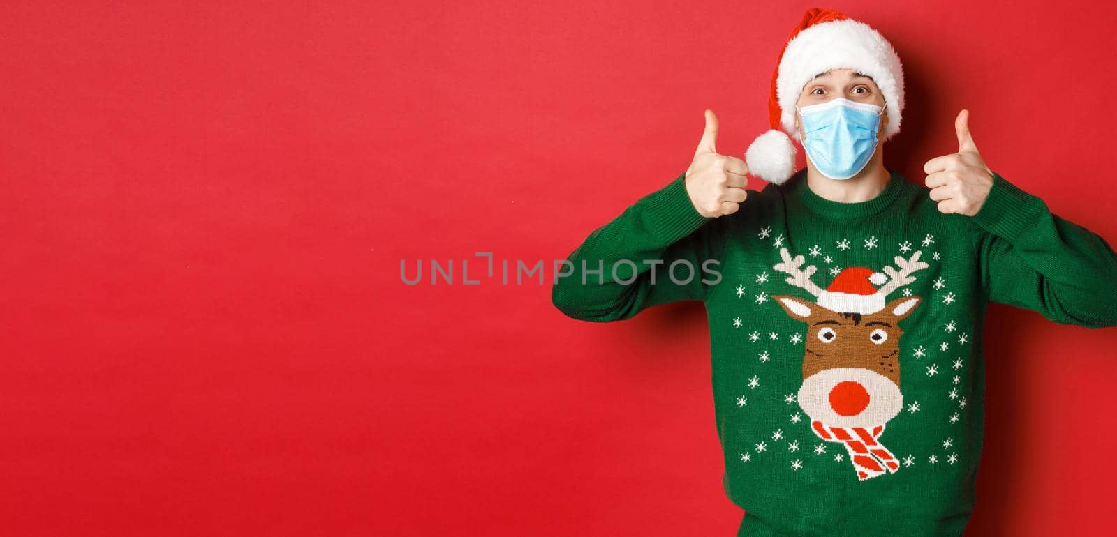 Concept of new year, covid-19 and social distancing. Cheerful handsome man in santa hat and medical mask, showing thumbs-up, enjoying christmas party, standing over red background.