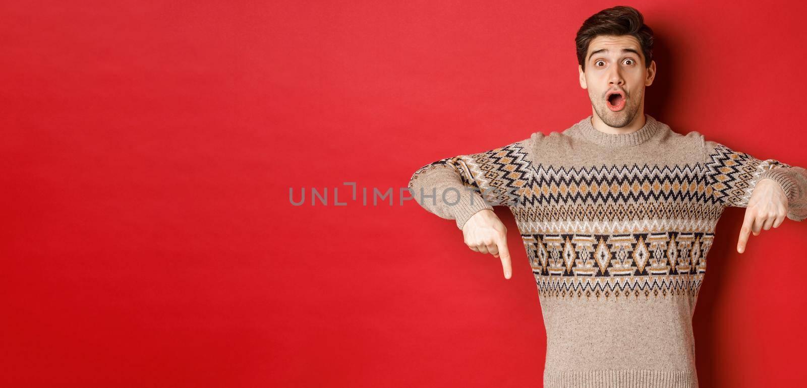 Concept of christmas celebration, winter holidays and lifestyle. Impressed handsome man in xmas sweater, pointing fingers down with amazed face, standing over red background.